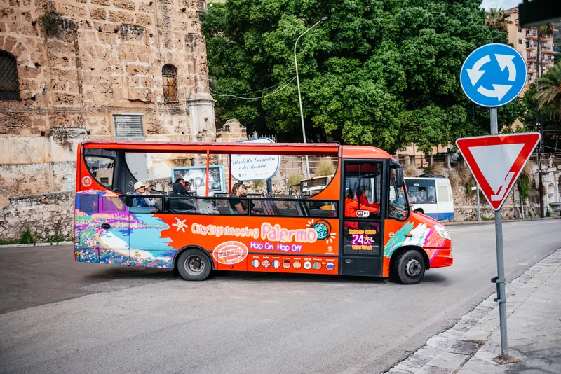 An open-top City Sightseeing Palermo hop-on hop-off bus parked on a street in Monreale beside the cathedral's stone wall