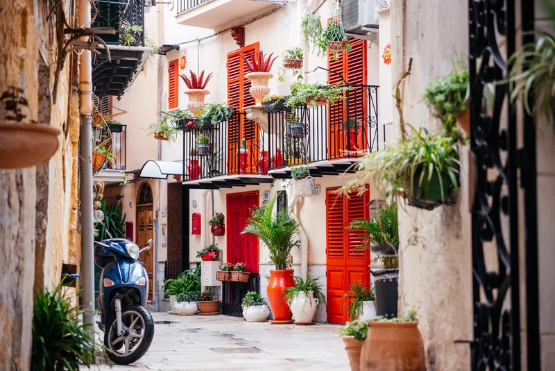 A narrow Monreale lane lined with orange shutters, potted plants and a parked scooter